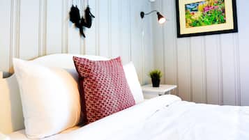 A neatly made bed with white and red pillows, a dreamcatcher, a framed picture, and a bedside lamp in a room with white paneled walls.