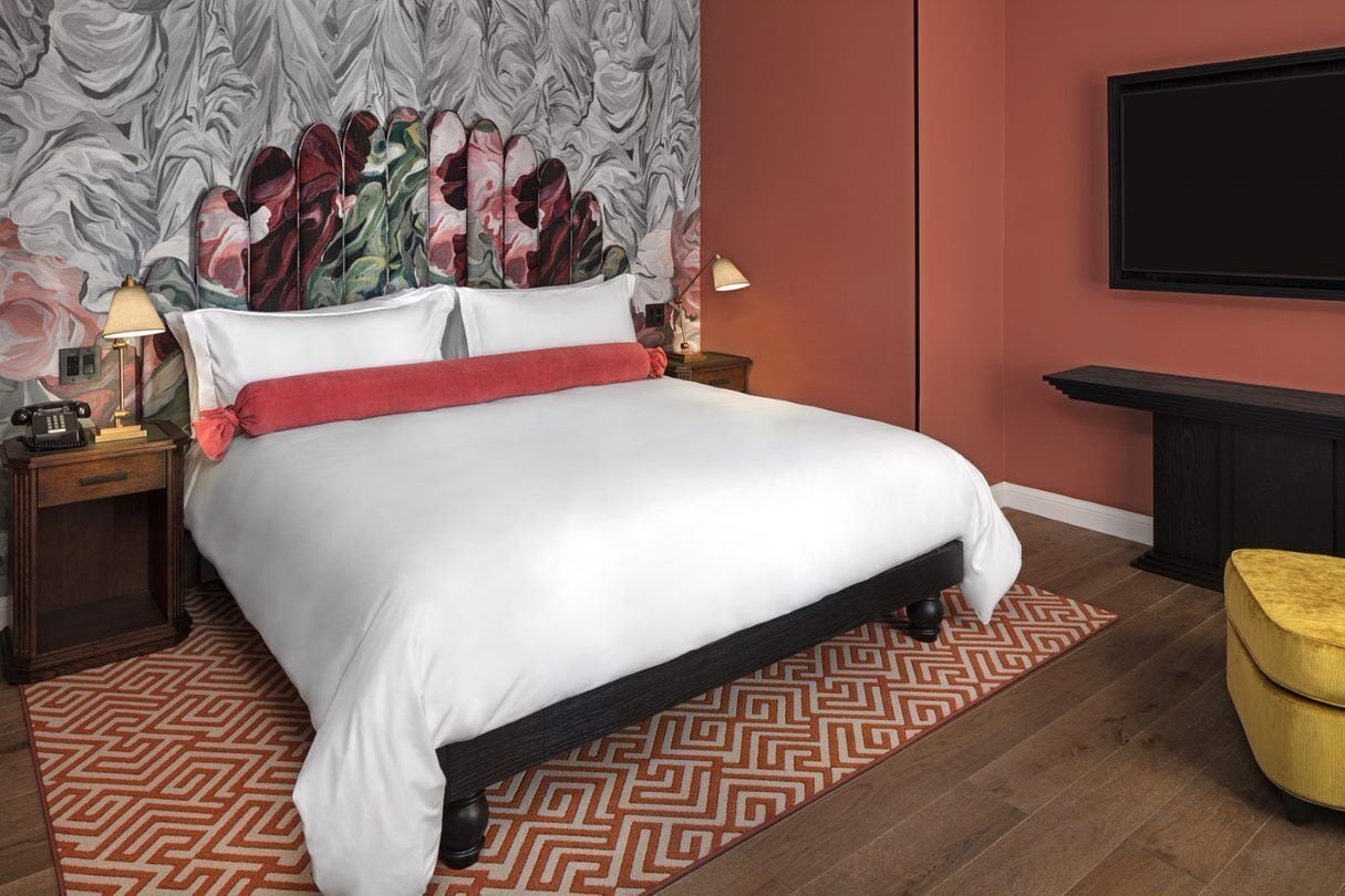 A neatly made bed with a red throw, a patterned rug, a headboard with a floral design, and a TV mounted on the wall.