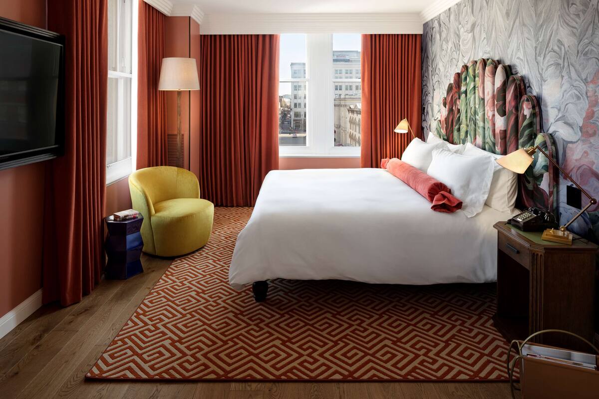 Habitación de hotel con una cama grande, un sillón amarillo, un televisor y vista a la ciudad por la ventana.