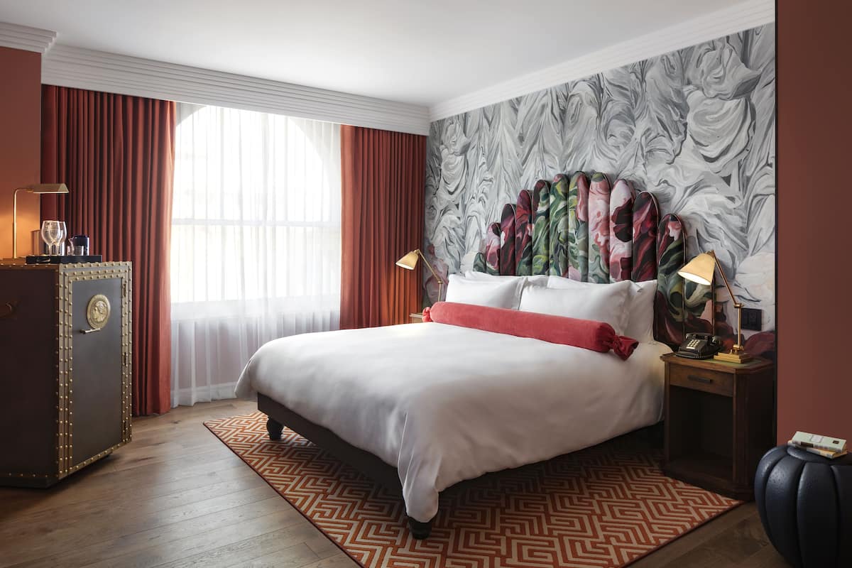 Una habitación de hotel con una cama grande, una mesita de noche con lámpara, un aparador y una ventana con cortinas.