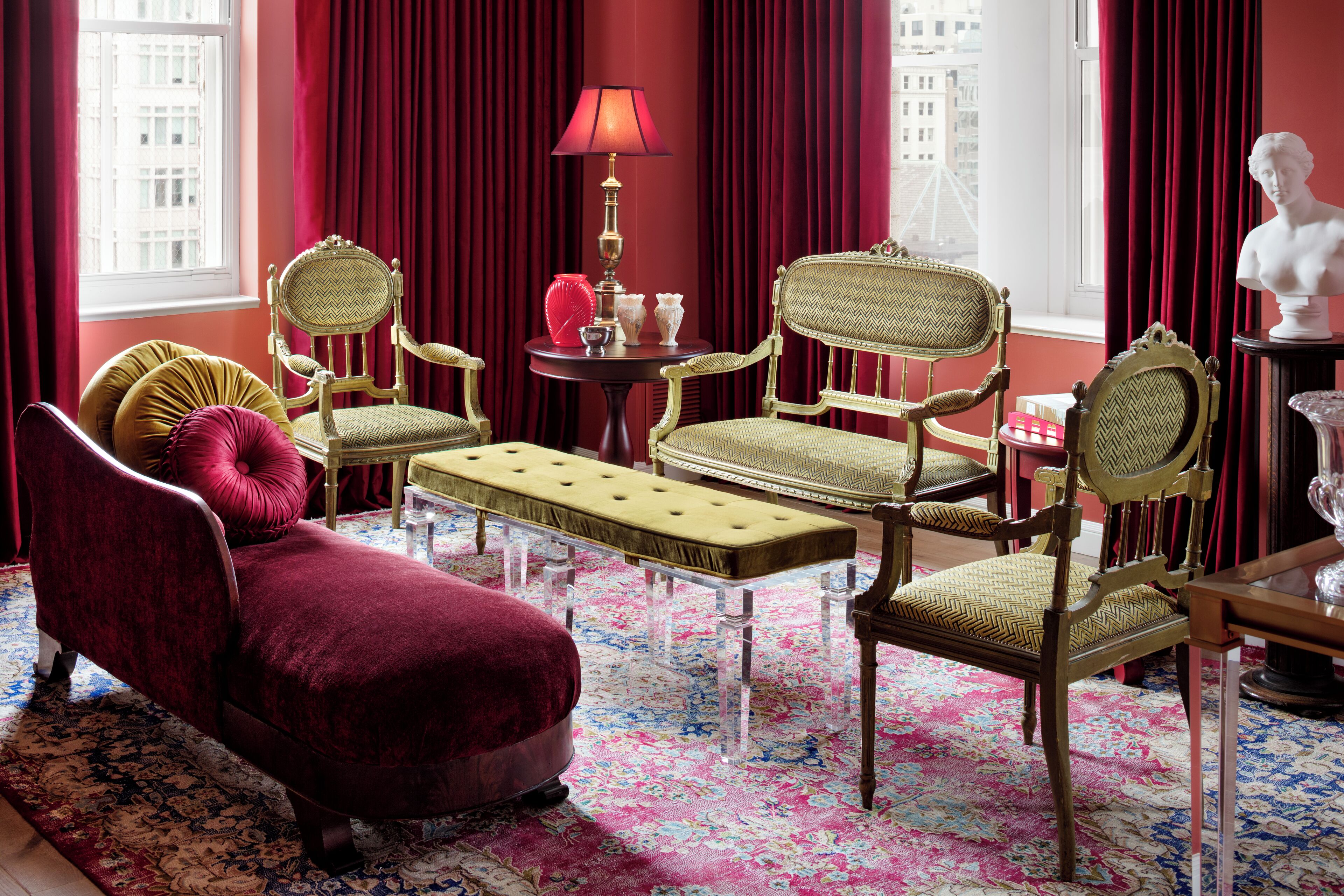 A room with a red velvet sofa, two green upholstered chairs, a small round table with a lamp, and a bust on a side table.
