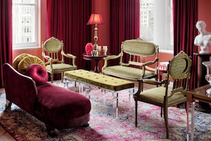 A room with a red velvet sofa, two green upholstered chairs, a small round table with a lamp, and a bust on a side table.
