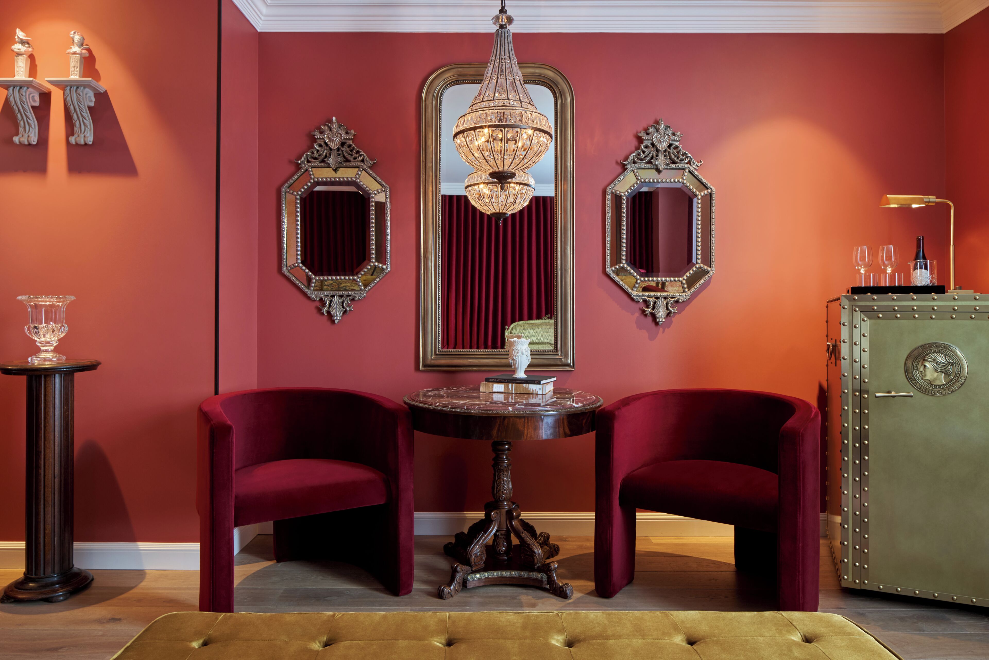 A room with two red velvet armchairs, a round table with a vase, and a green cabinet with a lock.