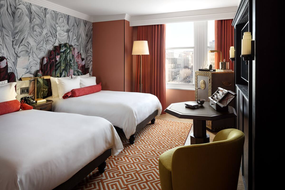 Habitación de hotel con dos camas, un escritorio con lámpara, una silla, un minibar y vistas a la ciudad.