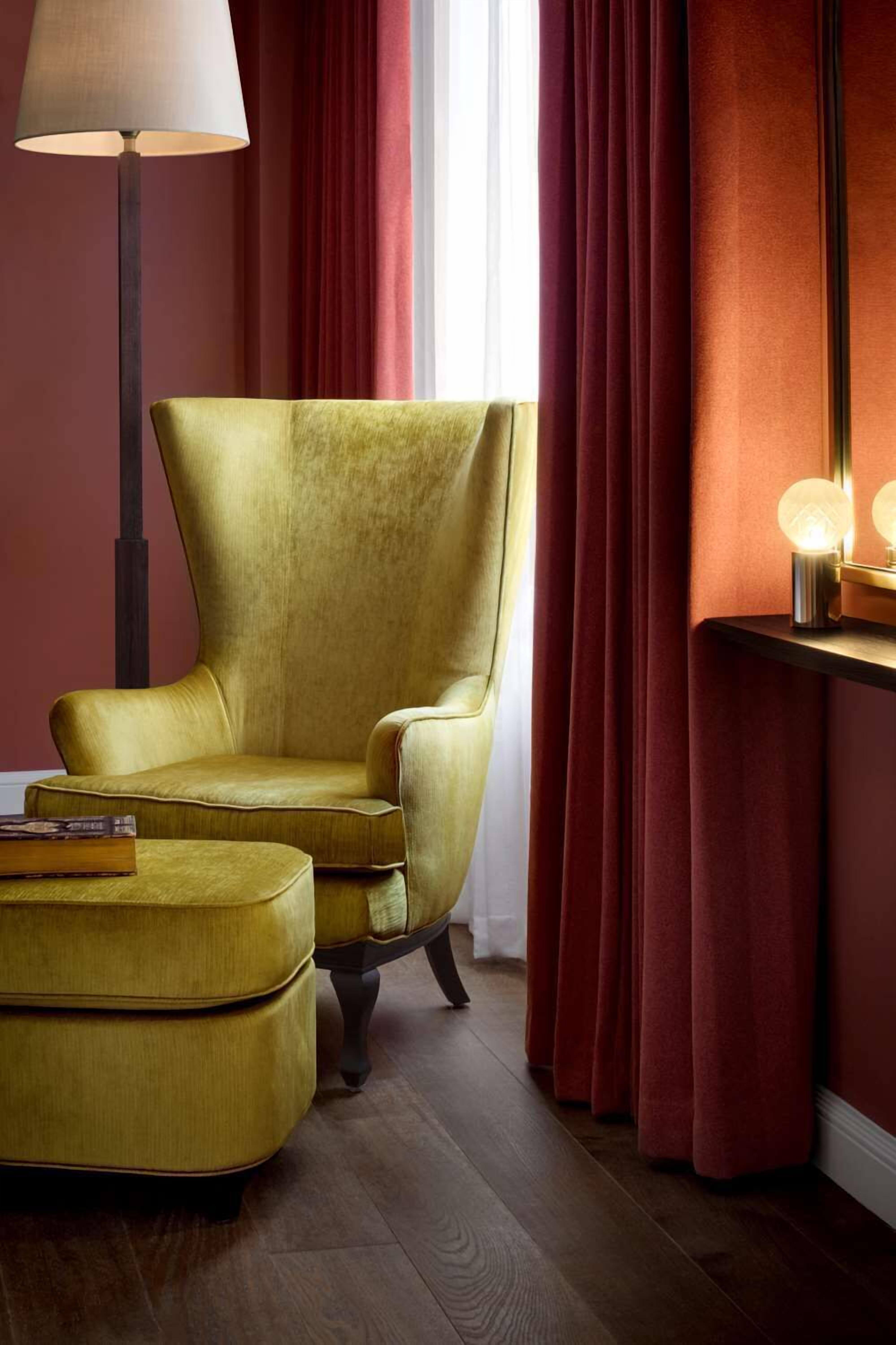 Een kamer met een grote, beklede fauteuil en een poef, een vloerlamp en een bijzettafeltje met een lamp.