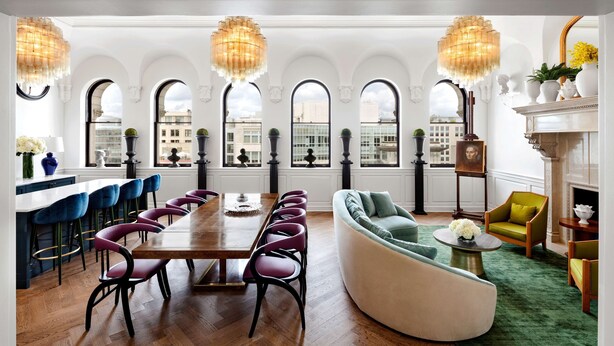A spacious dining area with a long wooden table and purple chairs, a curved sofa, and a fireplace with a green rug.