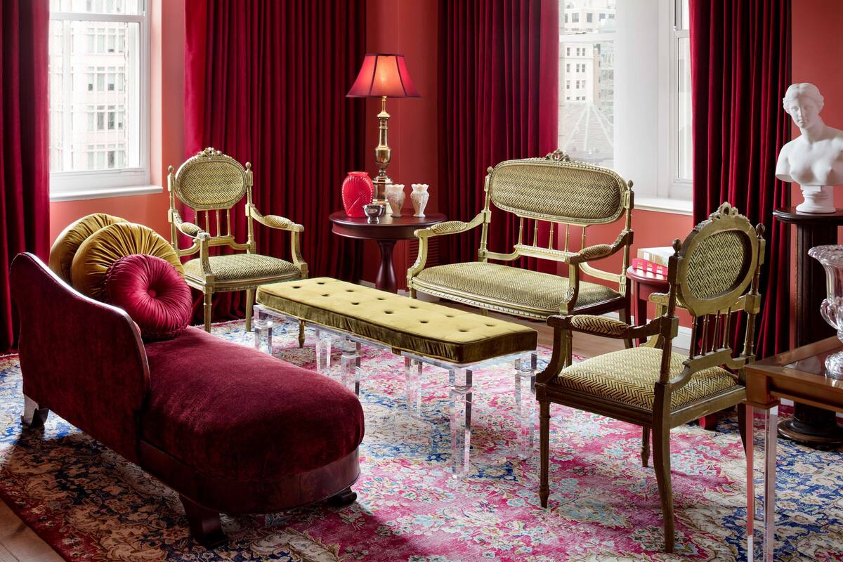 Una habitación con un sofá de terciopelo rojo, dos sillones de madera, una mesita redonda con una lámpara y un busto sobre una mesita auxiliar.