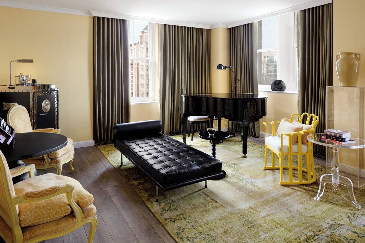 Una habitación con un sofá negro, un piano, sillas amarillas, una mesita y un amplio ventanal con cortinas.