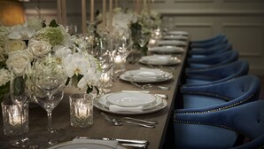 A long dining table set with white plates, silverware, and glassware, flanked by blue upholstered chairs.