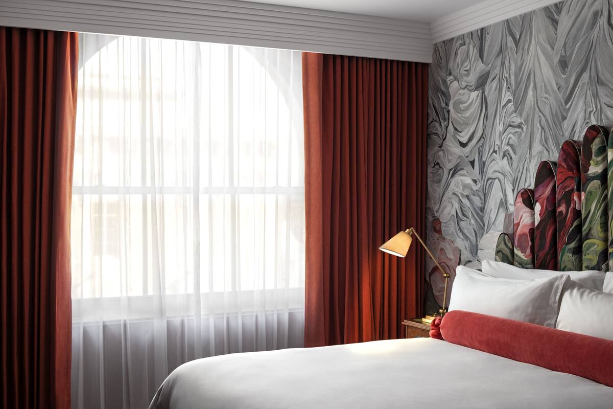Habitación de hotel con una cama grande, cortinas rojas, una pared con estampado floral y una lámpara en la mesita de noche.