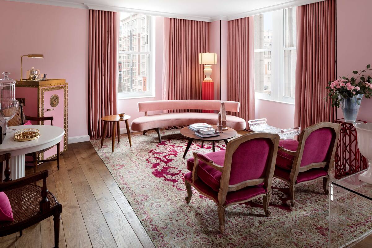 Una habitación con decoración rosa, que incluye una mesa de centro redonda, un sofá y dos sillones. Se observa una lámpara, un jarrón con flores y una ventana con cortinas.