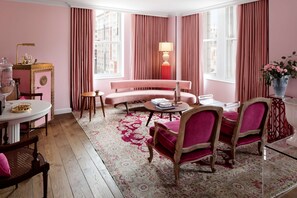 A pink-themed room with a round coffee table, a sofa, and two armchairs. There is a lamp, a vase with flowers, and a window with curtains.