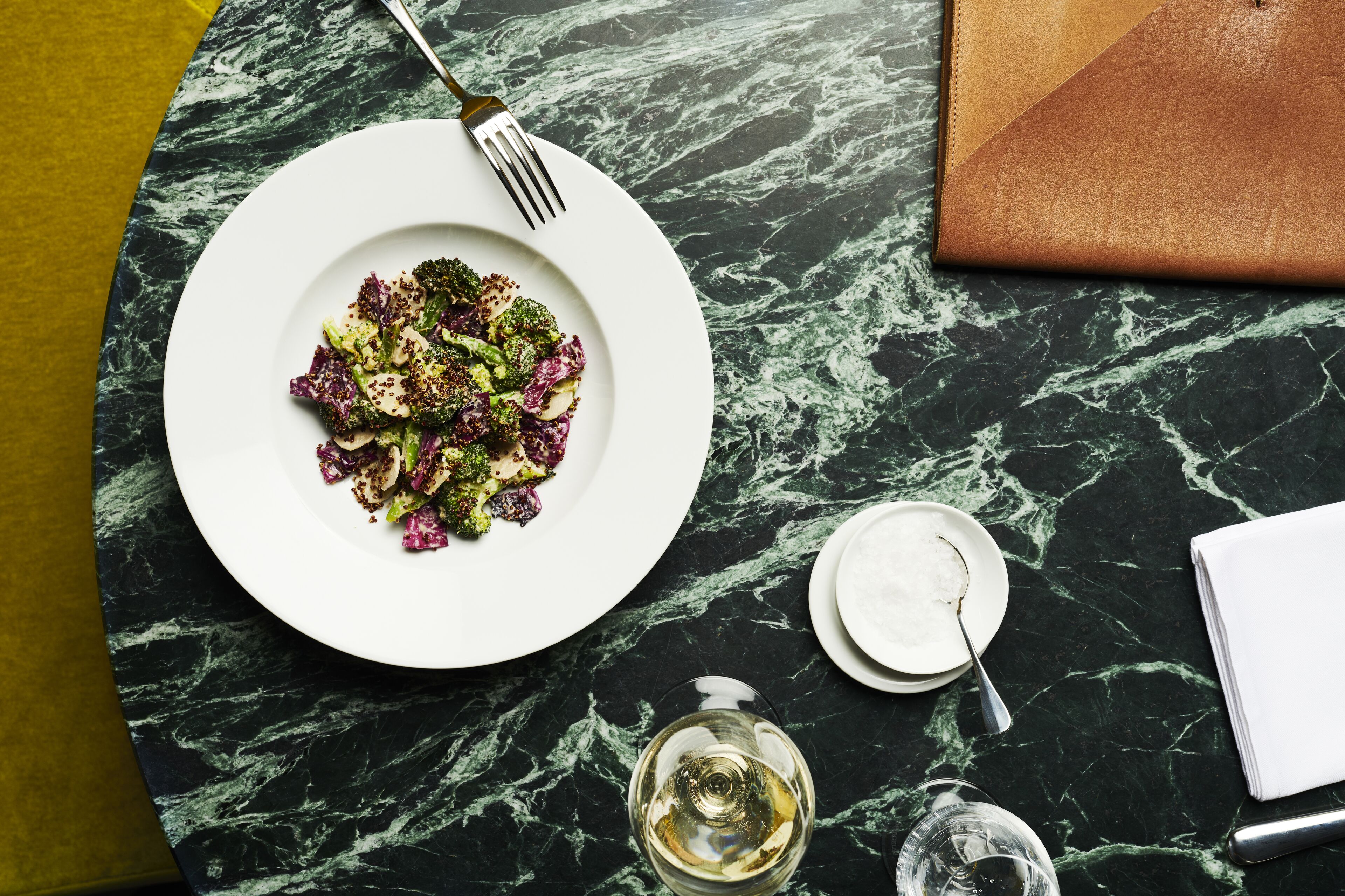 Une table à manger avec une assiette de nourriture, une fourchette, un verre de vin blanc et une serviette.