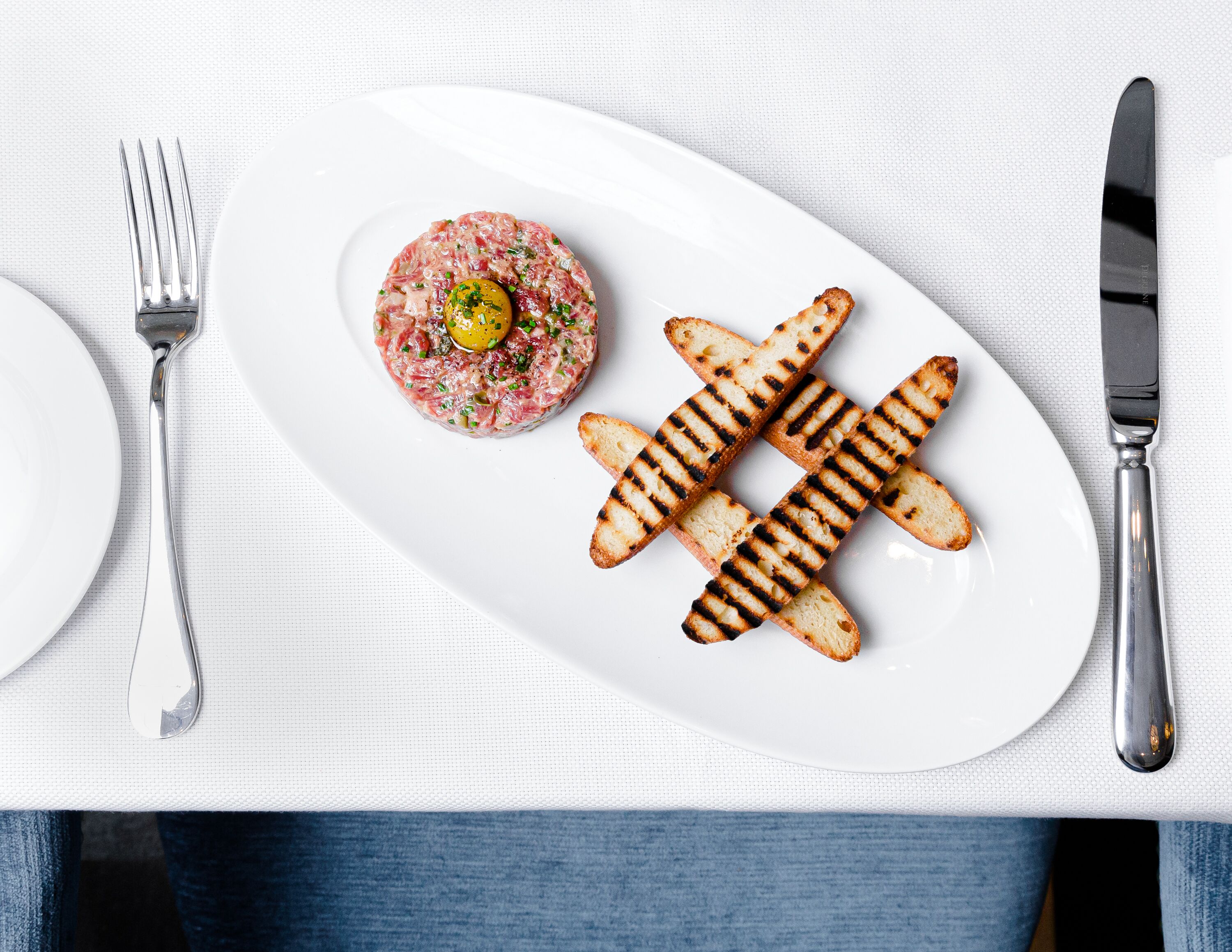 Un piatto bianco con tartare di manzo e pane grigliato su una tovaglia bianca.