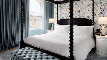 A four-poster bed with white linens, a tufted ottoman, and a nightstand with a lamp. The room has a patterned wallpaper and a view of a building through a window with blue curtains.
