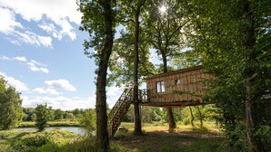 A treehouse with a wooden exterior, a staircase leading up to it, surrounded by lush greenery and a pond. - Camping Les Castors - Cabanes Insolites (Burnhaupt-le-Haut)
