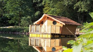 A wooden birdhouse with a balcony, situated by a calm lake surrounded by dense greenery. - Camping Les Castors - Cabanes Insolites (Burnhaupt-le-Haut)