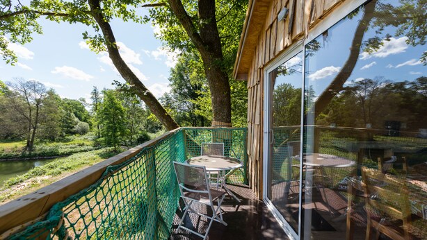 A glass-enclosed outdoor patio with a table and chairs, surrounded by a green fence, overlooking a river and trees. - Camping Les Castors - Cabanes Insolites (Burnhaupt-le-Haut)