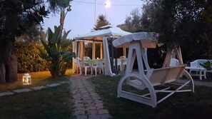 An outdoor seating area with white plastic chairs and a wooden swing chair, surrounded by greenery and a paved path. - Residence Alba (Vieste)