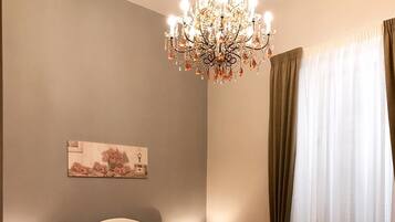 A bedroom with a large bed, a chandelier, a bedside table with a lamp, a painting on the wall, and a window with curtains.
