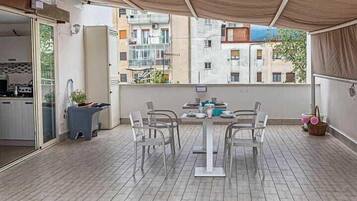Un balcon avec une table et des chaises, une porte-fenêtre coulissante donnant sur une cuisine et une vue sur les immeubles voisins.