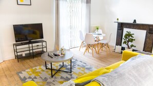 Een moderne woonkamer met een gele bank, een salontafel, een tv op een meubelstuk, een open haard en een eethoek met stoelen.