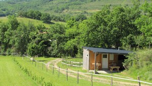 A small wooden cabin with a metal roof, surrounded by a fenced area and lush greenery. - Camping des vignes (Dun)
