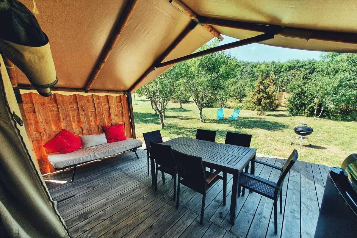 Una terraza exterior con mesa y sillas, un sofá con cojines rojos y una parrilla de barbacoa en un área boscosa.