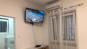 A bedroom with a bed, a TV mounted on the wall, a desk with chairs, a window with a patterned curtain, and a bathroom with a toilet and a towel dispenser. - Rooms Radunica street (Split)