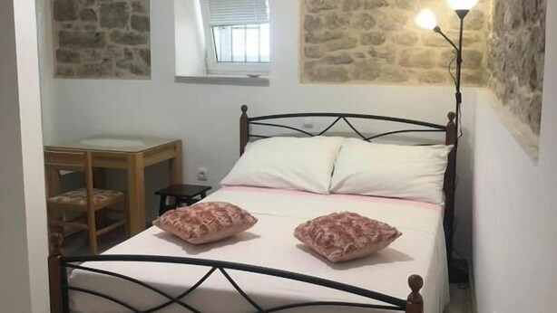 A bedroom with a stone wall, a bed with a metal frame, a bedside table, a chair, and a lamp. - Rooms Radunica street (Split)