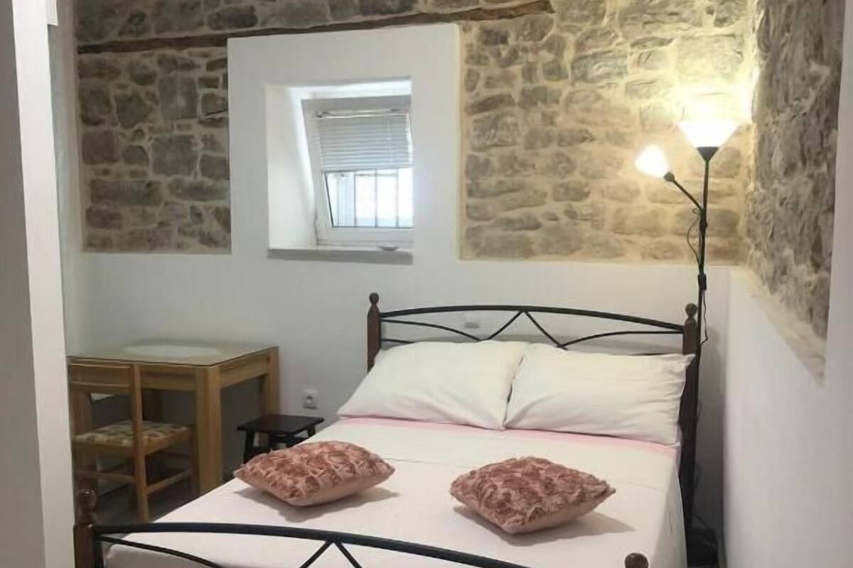 A bedroom with a stone wall, a bed with a metal frame, a bedside table, a chair, and a lamp.