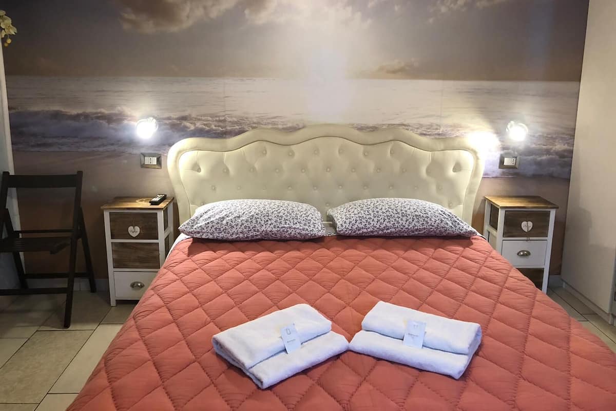 A bedroom with a bed, two nightstands, a chair, and a wall mural of a cloudy sky over water.