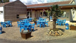 A building with a tiled roof, blue plastic chairs, and a small palm tree. - Camping La Croix Badeau Tentes équipées (Soulaines-Dhuys)