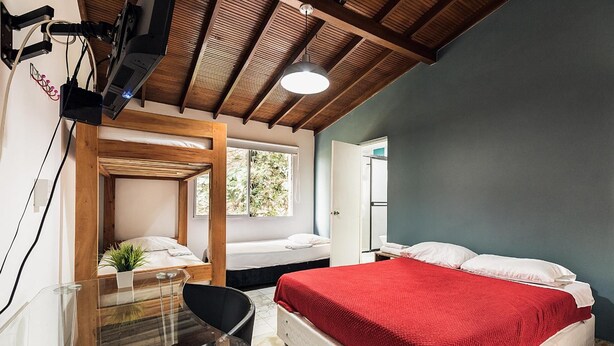 A modern bedroom with a wooden ceiling, a glass table, a bed with a red blanket, and a window with a view of greenery. - Living Hostal - Hostel (Medellín)
