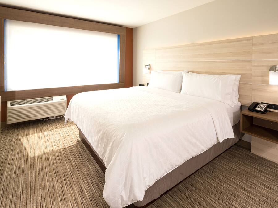 A hotel room with a large bed, a bedside table with a phone, a wall-mounted light, and a large window with blinds.