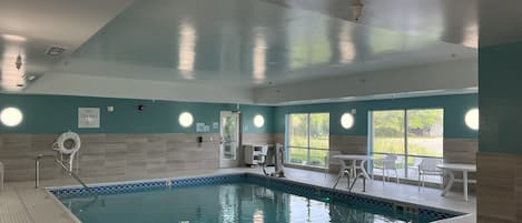 Una piscina interior con agua cristalina, rodeada de un suelo y paredes de baldosas. Hay sillones de descanso y una mesa cerca de la piscina, y grandes ventanales que dejan entrar luz natural.