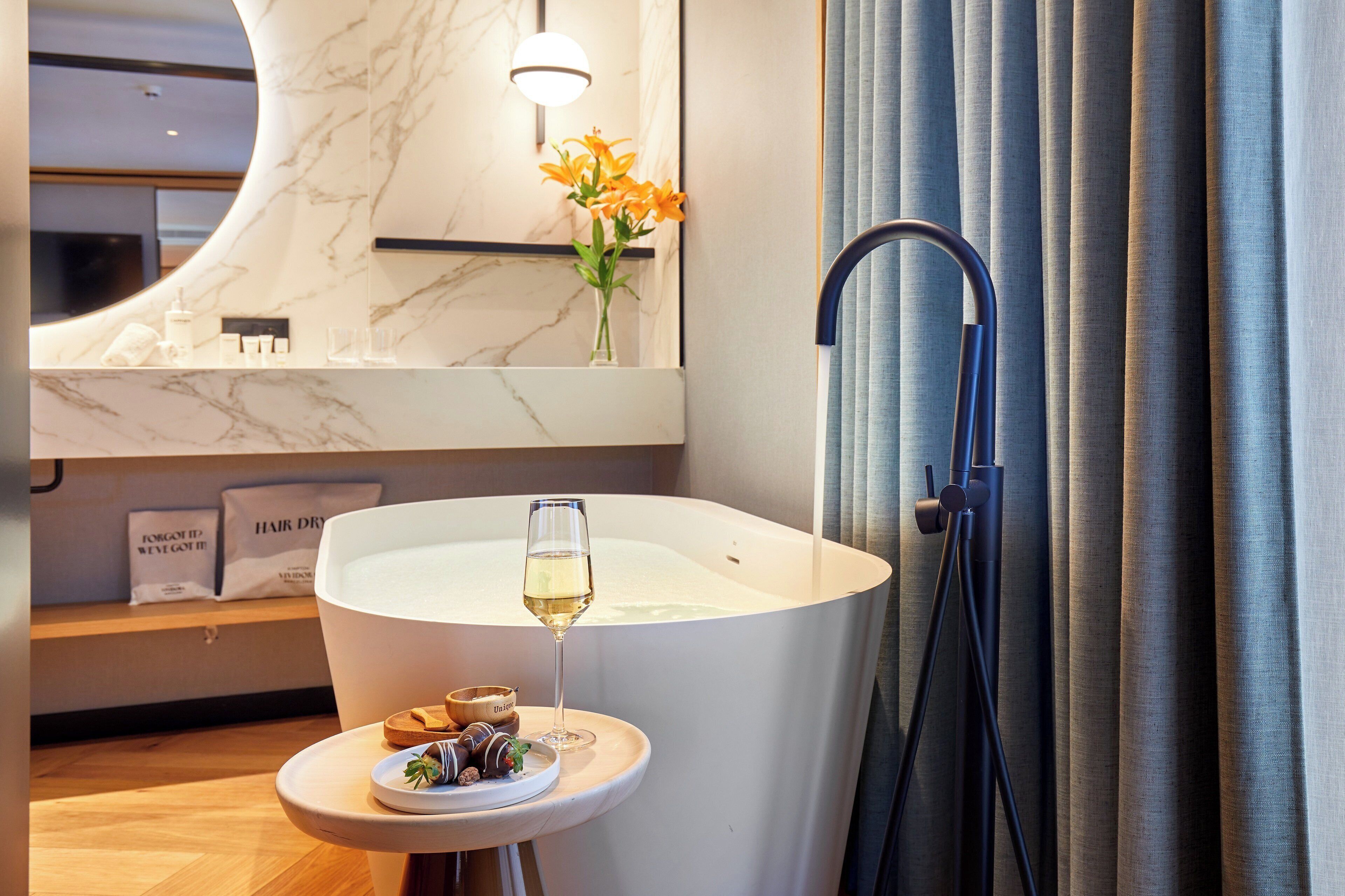 A modern bathroom with a large bathtub, a round mirror, and a small table with a glass of wine and chocolates.