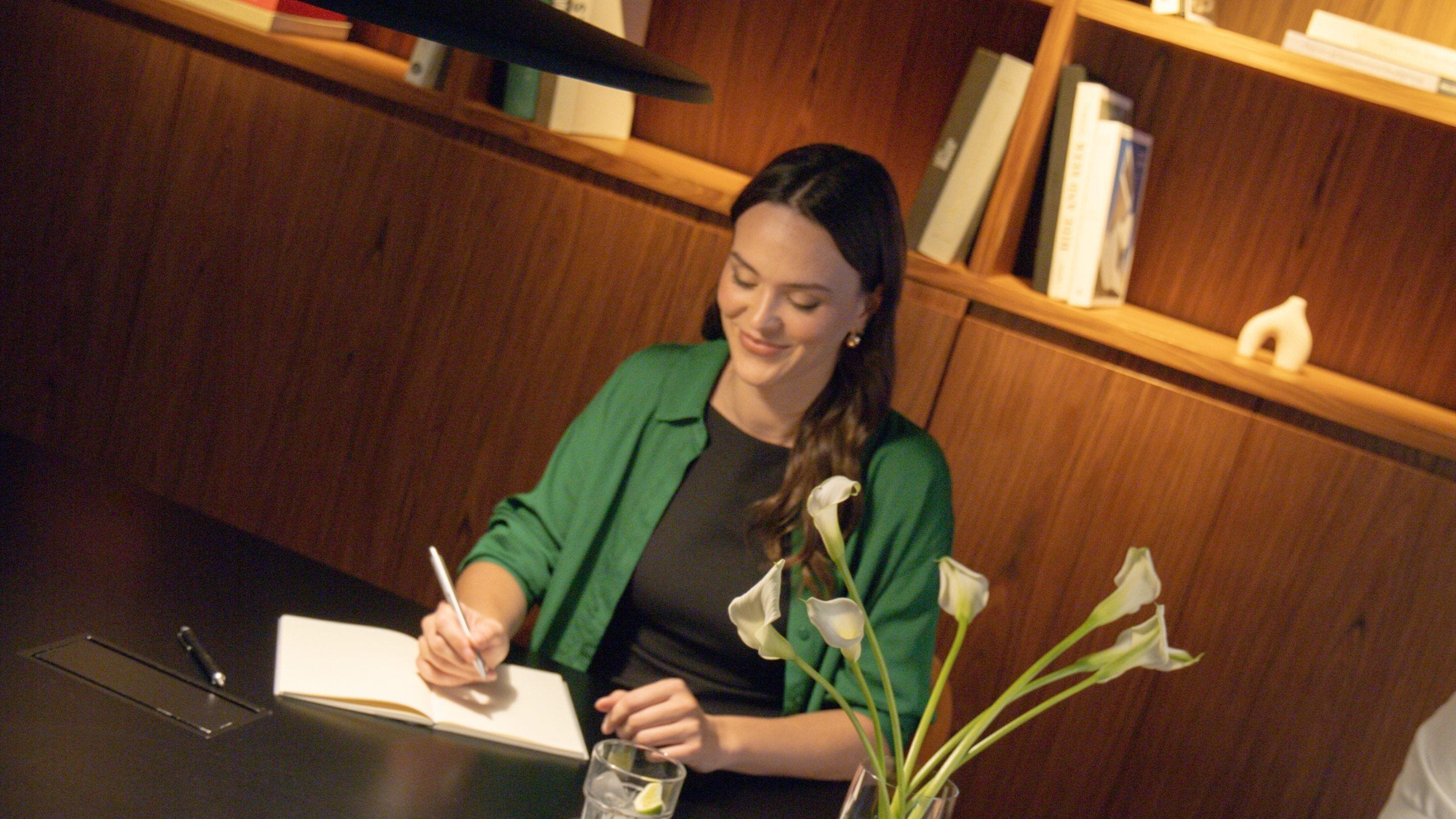 A person sitting at a table with a book, writing, and a vase with flowers.