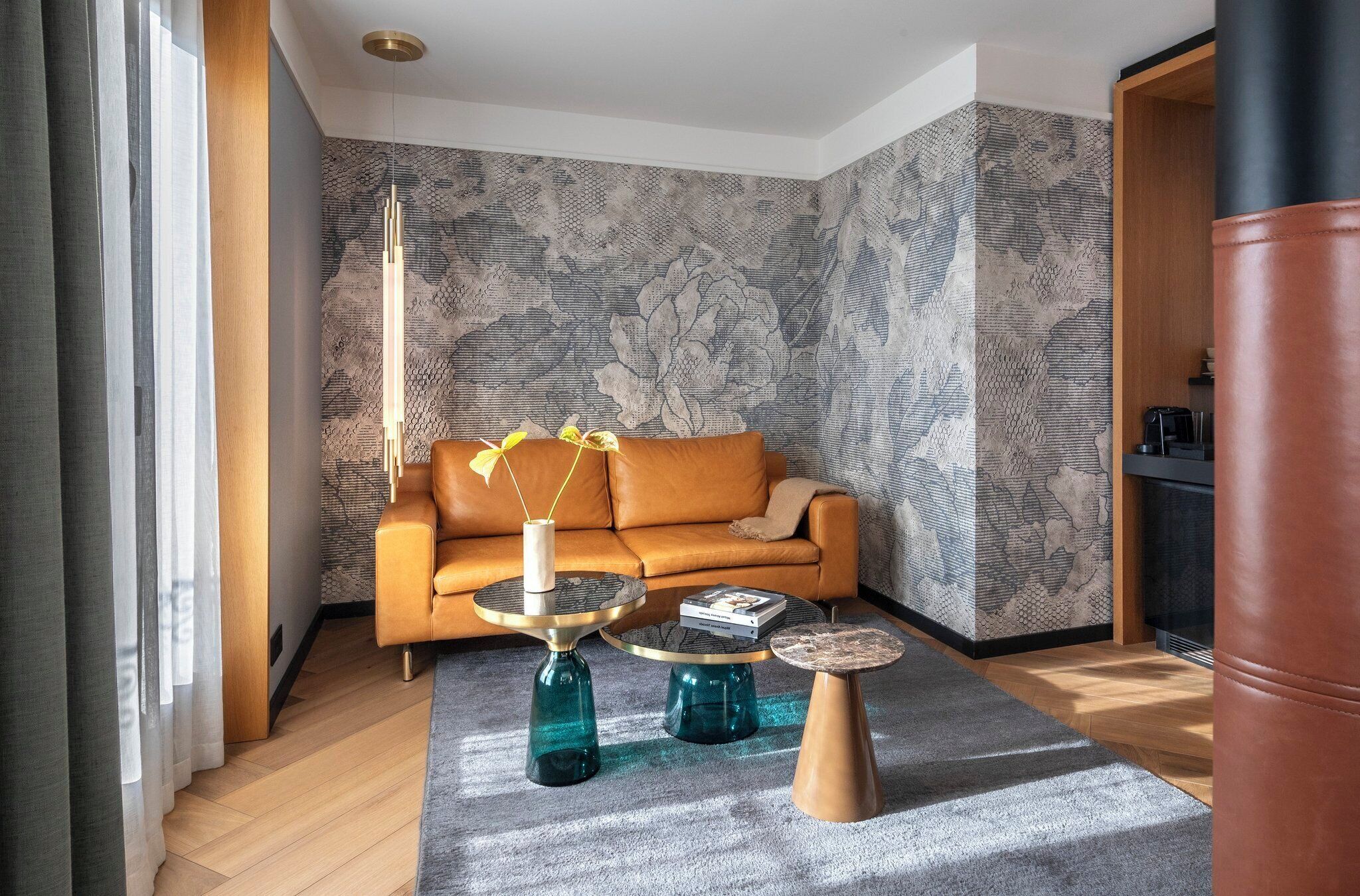 Een moderne woonkamer met een leren bank, drie glazen salontafels en behang met een patroon.