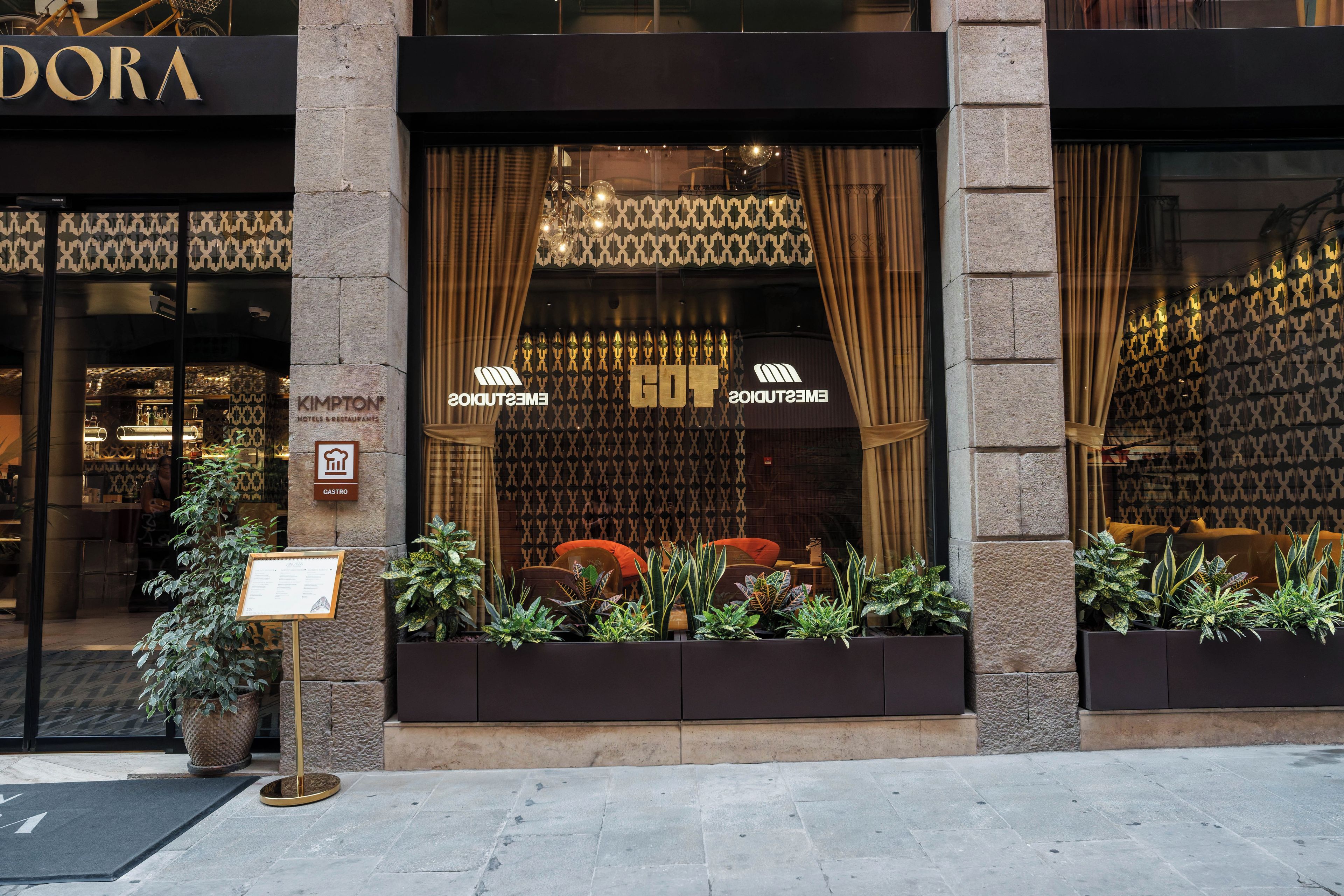 A hotel entrance with a sign for 'Gastro' indicating a restaurant. There are potted plants in front of the entrance.