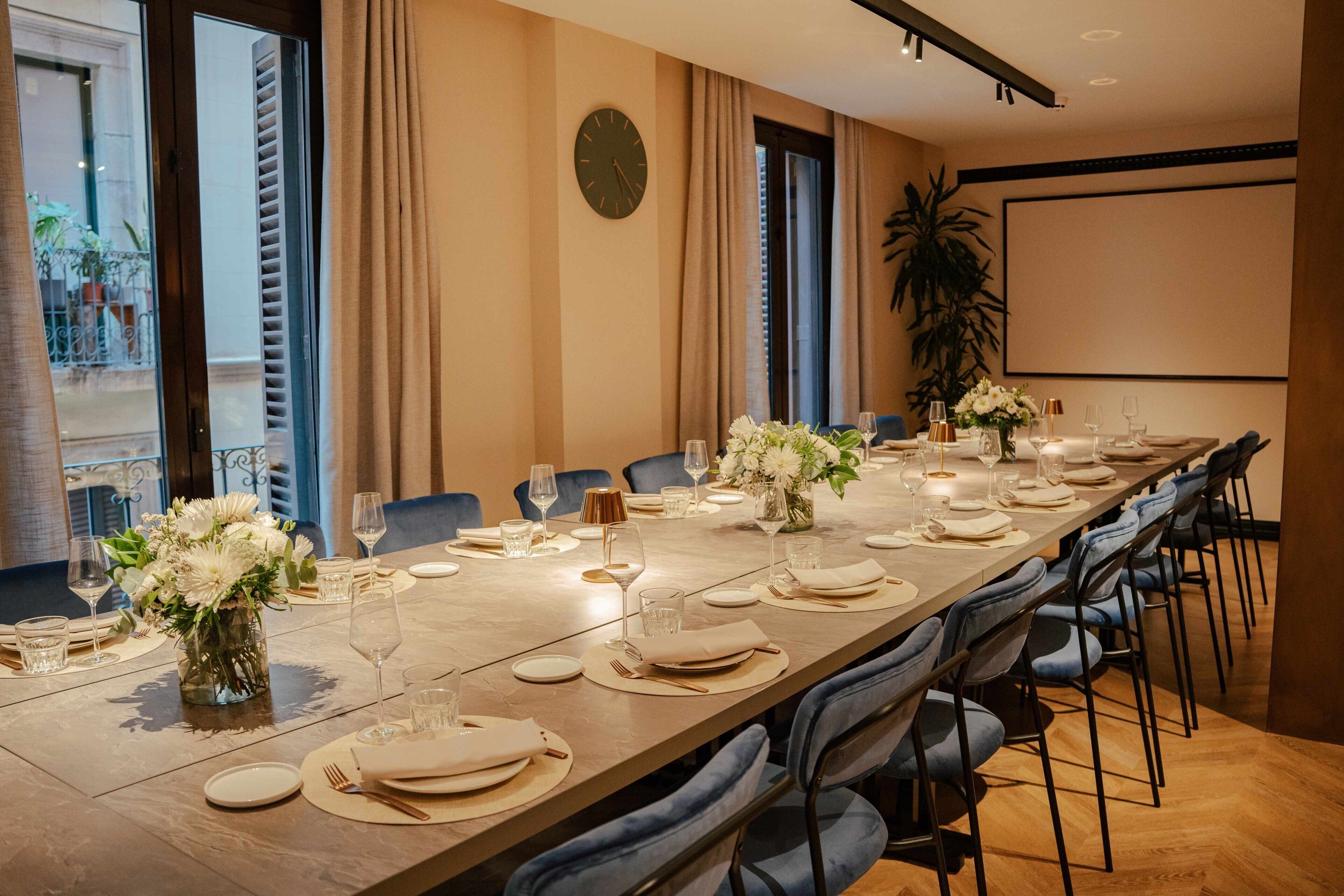 Una larga mesa de comedor dispuesta para una comida formal, con sillas, un reloj y una planta grande al fondo.