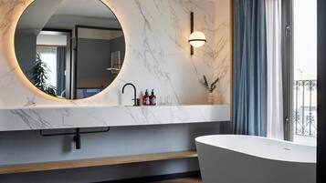 Un baño moderno con tocador de mármol, espejo redondo, bañera independiente y vista a la ciudad por una ventana.