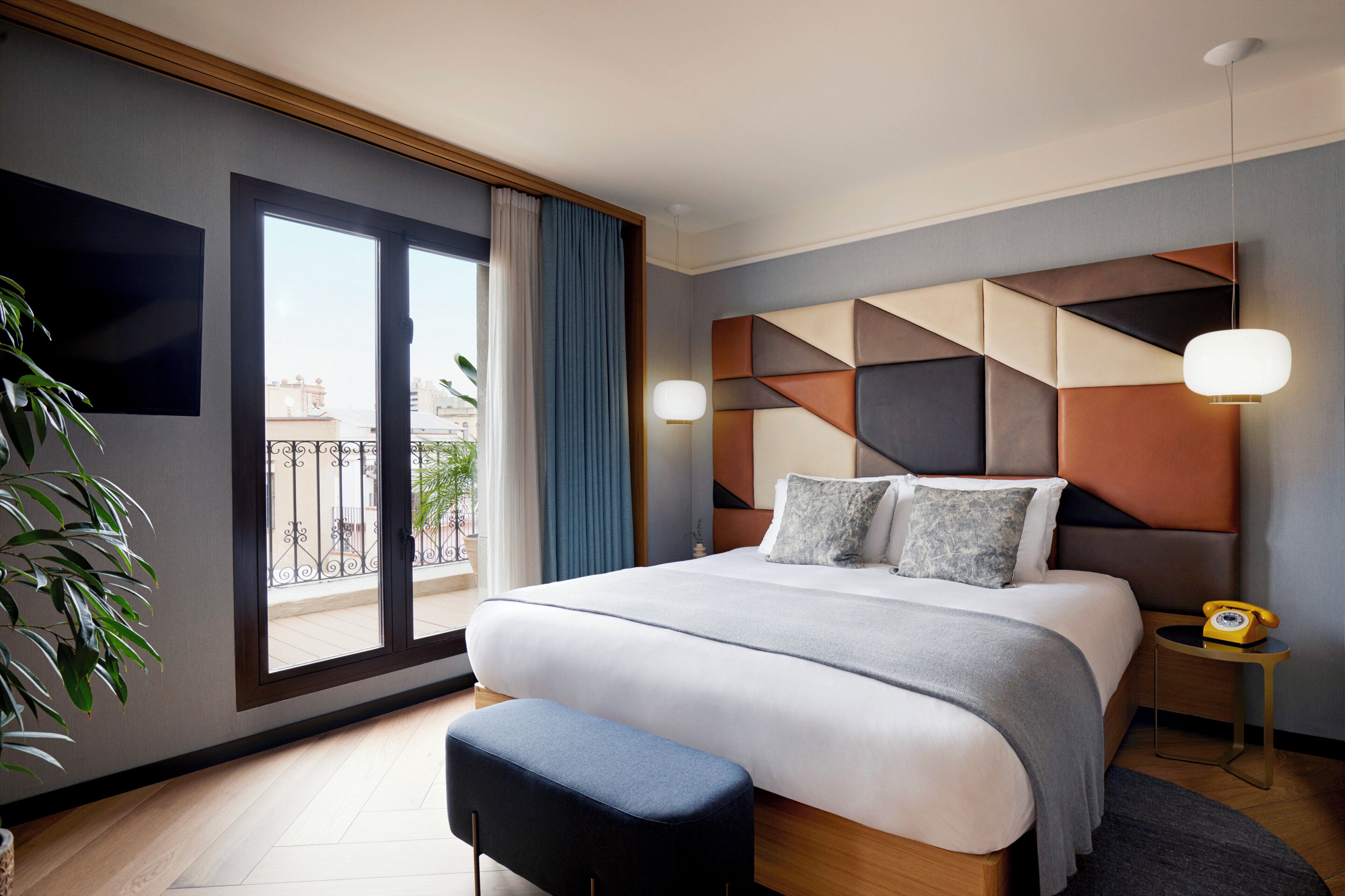 A modern hotel room with a large bed, a small bedside table, a nightstand, a potted plant, and a view of the city through the window.