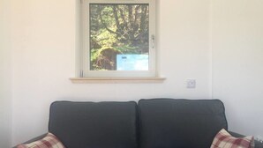A dark-colored sofa with two plaid cushions, a window with a view of a forest and a blue structure, and a light switch on the wall. - Cosy sea View Cottage Isle of Skye (Glendale)