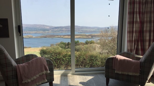 A room with two plaid armchairs facing a large sliding glass door leading to a scenic outdoor view. - Cosy sea View Cottage Isle of Skye (Glendale)