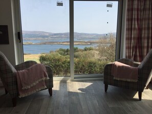 A room with two plaid armchairs facing a large sliding glass door leading to a scenic outdoor view. - Cosy sea View Cottage Isle of Skye (Glendale)