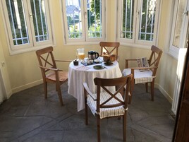 Un comedor con una mesa redonda, cuatro sillas de madera, un mantel blanco y comida para el desayuno.
