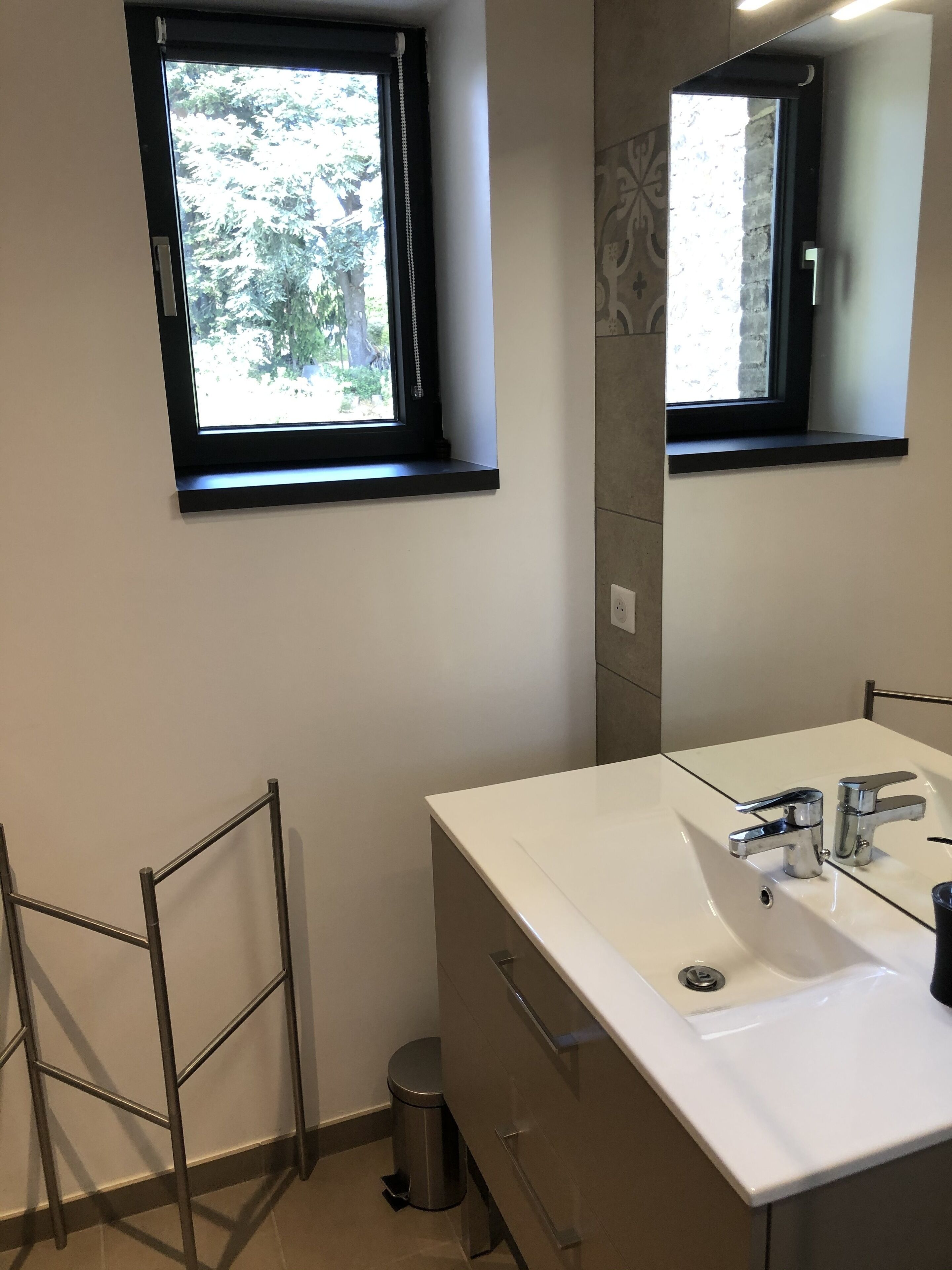 Ein Badezimmer mit einem weißen Waschbecken, zwei Wasserhähnen, einem Spiegel und einem Fenster mit Blick auf Bäume.