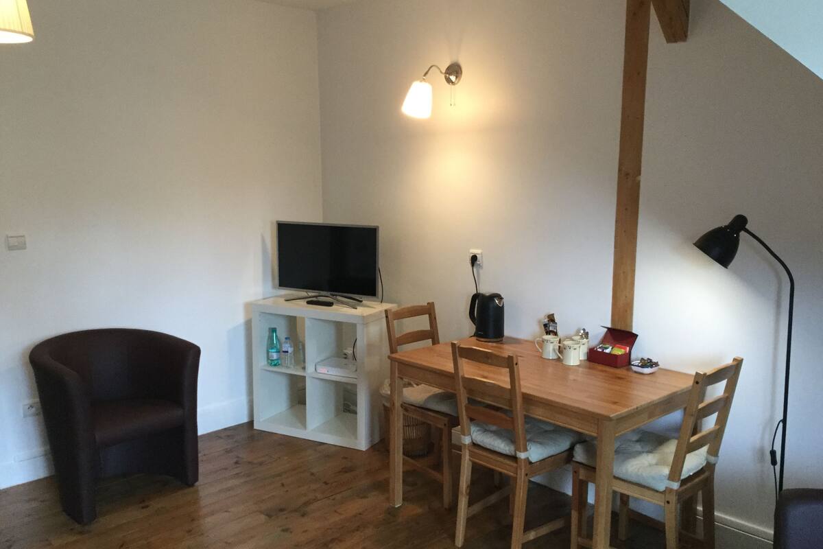 Een eethoek met een houten tafel en stoelen, een televisie op een witte kast, een koffiezetapparaat en een lamp.