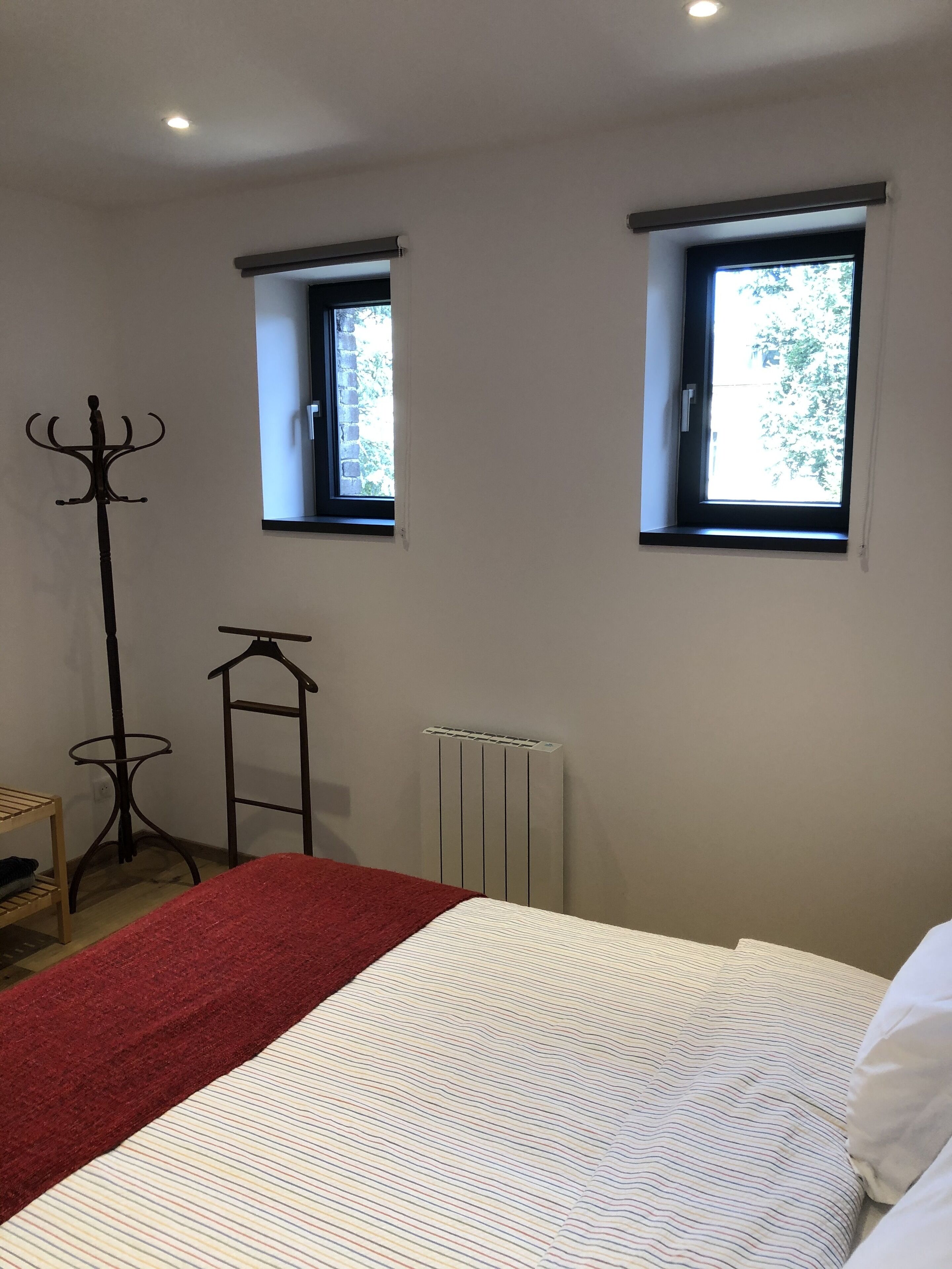 A bedroom with a bed, two windows, a radiator, and a coat rack.